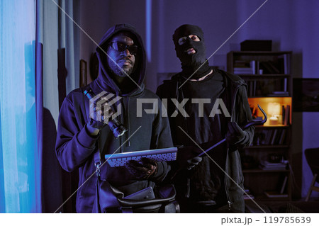 Two thieve in black clothes with crowbar, flashlight and stolen keyboard in hands hiding his face and head under hood and balaclava posing for portrait in dark room 119785639