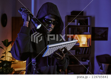 African American in total black clothes with hood looking at wireless keyboard in dark room 119785641