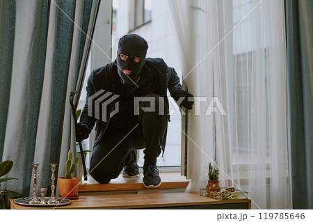 Man in total black with balaclava and crowbar sitting on windowsill after break-in Man in total black with balaclava and crowbar sitting on windowsill after break-in 119785646