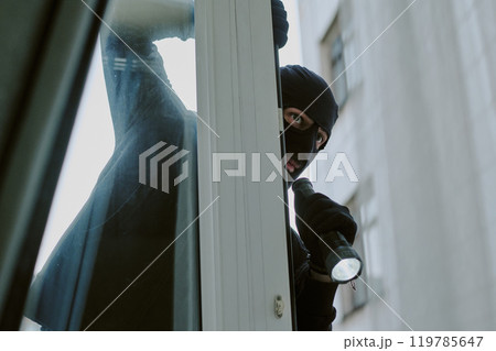 Low angle shot of unrecognizable thief in balaclava with flashlight seeing apartment from window 119785647