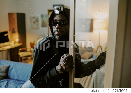 African American thief with headlamp opening cupboard carefully while glancing around 119785661