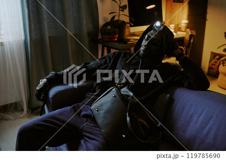 African American thief in black clothes and leather gloves sitting on sofa relaxed during trespassing 119785690