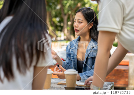 An Asian female college student is discussing work with her friends at a table in a college park. An Asian female college student is discussing work with her friends at a table in a college park. 119785733