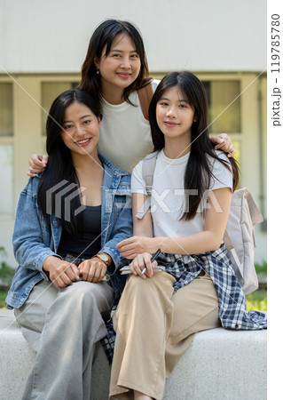 A group of Asian students is sitting on a bench in their college park, smiling at the camera. A group of Asian students is sitting on a bench in their college park, smiling at the camera. 119785780