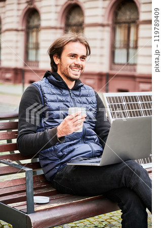 Man sits on wooden bench in urban setting, working on laptop with cup of coffee in hand. Solar panel rests beside, emphasizing sustainable, mobile workspace amidst backdrop of historic architecture. Man sits on wooden bench in urban setting, working on laptop with cup of coffee in hand. Solar panel rests beside, emphasizing sustainable, mobile workspace amidst backdrop of historic architecture. 119789409