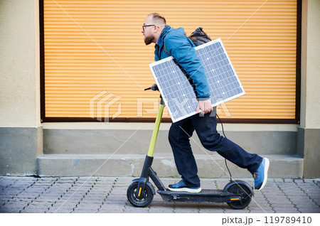 Man rides electric scooter while carrying solar panel. Integration of solar power as sustainable energy source for charging electric scooters, promoting eco-friendly urban transportation. 119789410