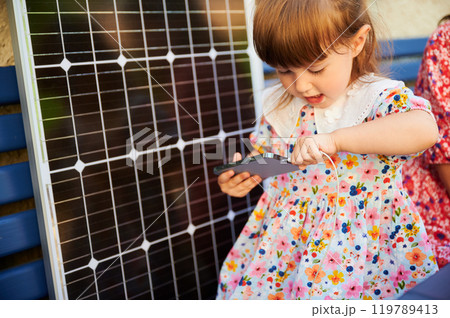 Young girl holds smartphone connected to solar panel for charging. Integration of sustainable renewable energy into daily life, importance of teaching children about eco-friendly technology. 119789413