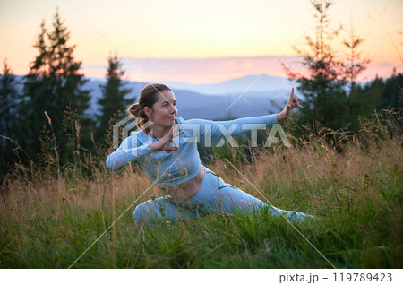 Woman practicing yoga outdoors in the mountains in a serene, natural setting. Female performing yoga pose, with backdrop of beautiful mountain landscape at sunrise or sunset. 119789423