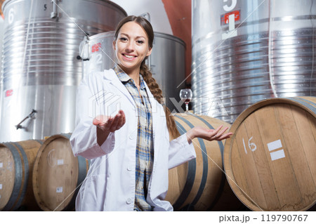 Specialist in white coat examines glass of wine on the background of barrels for fermentation 119790767