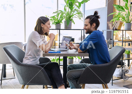 Young man woman sitting at table in coworking space with laptop smartphone, talking together Young man woman sitting at table in coworking space with laptop smartphone, talking together 119791551