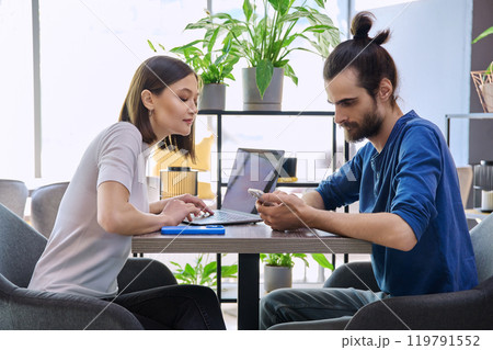 Young man woman sitting at table in coworking space with laptop smartphone, talking together Young man woman sitting at table in coworking space with laptop smartphone, talking together 119791552