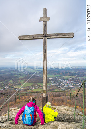 Two hikers rest by the wooden summit cross on Oresnik granite rock formation, taking in the panoramic views of Hejnice and the Jizera Mountains in Czechia. 119795741