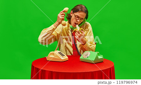 Man stares at banana while talking on two vintage phones, adding quirky and humorous twist to office scene. 119796974