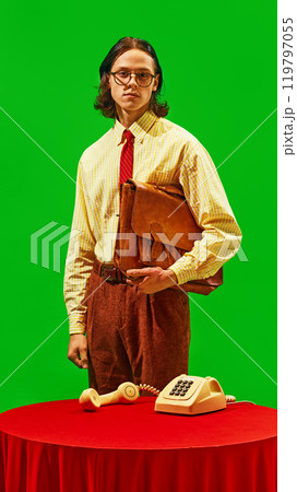 Young man in vintage attire holds briefcase, against green background, and stands next to old-fashioned rotary phone. Retro office environment Young man in vintage attire holds briefcase, against green background, and stands next to old-fashioned rotary phone. Retro office environment 119797055