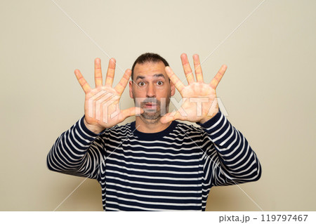 Bearded hispanic man in his forties wearing a striped sweater feels dumbfounded and scared, fearing something scary, with open hands in front. Isolated over beige studio background 119797467