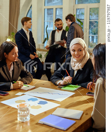 Multicultural female colleagues engaged in discussion during corporate strategy meeting. Other team, male members talking in the background. 119797987
