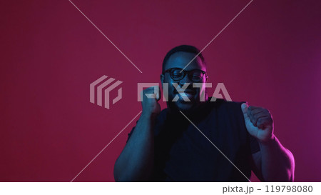 Winner dance. Victory celebration. Overjoyed funky excited smiling man satisfied with success in neon light isolated on dark red background empty space. 119798080