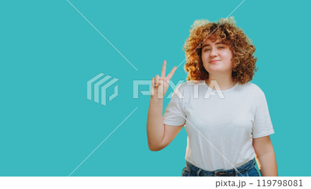 Peace sign. Positive emotion. Joyful happy smiling overweight girl with curly hair showing victory hand gesture isolated on blue empty space background. 119798081