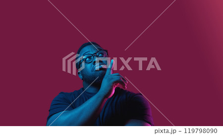 Pointing advertisement. Product promotion. Casual doubtful man in neon light demonstrating offer with finger up isolated on burgundy background empty space. 119798090