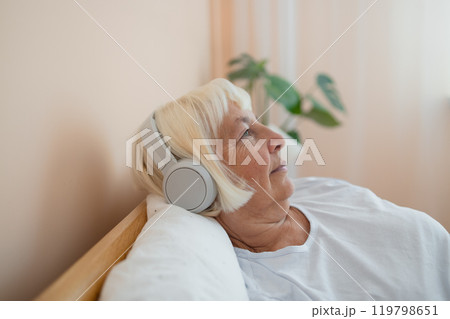 Calm senior woman in headphones enjoying audiobook, resting in bed in the morning Calm senior woman in headphones enjoying audiobook, resting in bed in the morning 119798651