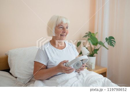 Calm senior woman in headphones enjoying audiobook, resting in bed in the morning Calm senior woman in headphones enjoying audiobook, resting in bed in the morning 119798652