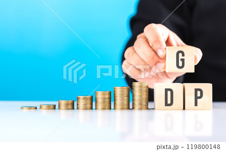 Hand holding a wooden block labeled 'GDP,' with a stack of coins placed on a table. Symbolizes economic growth, highlighting the role of Gross Domestic Product in measuring financial prosperity Hand holding a wooden block labeled 'GDP,' with a stack of coins placed on a table. Symbolizes economic growth, highlighting the role of Gross Domestic Product in measuring financial prosperity 119800148