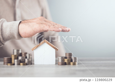 Businessman making a protective hand gesture over a stack of coins and a house model. Image represents the concept of asset protection, emphasizing safeguarding financial investments and property 119800229