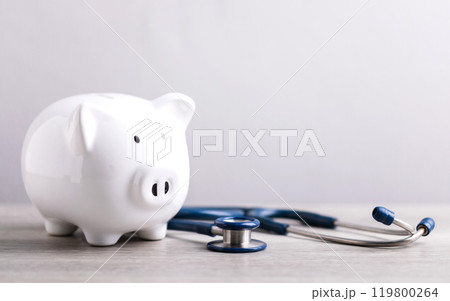 A white piggy bank and a stethoscope rest on a table against a bright white background. The image represents the concept of saving money for health care or financial security. 119800264
