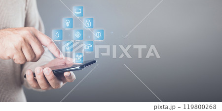 A man operates his smartphone to control smart home systems via the internet.  Home security icons highlight the advanced safety features, showcasing the ease and efficiency of modern home automation 119800268