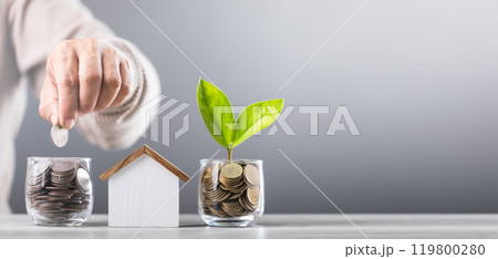 Man drops coins into a glass jar and a  wooden house model beside him. Emphasizes the concept of saving money for future house investments and financial stability 119800280