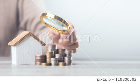 Mand holding a magnifying glass over a stack of coins and a house model. The image represents assessing home damage and budgeting for repairs, emphasizing careful financial planning for maintenance 119800302