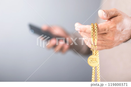 A close-up of a man holding a gold necklace in his left hand while checking his phone with his right hand. Concept of monitoring gold prices, symbolizing market speculation and investment trends 119800371