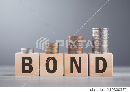 A hand stacking coins on a wooden block labeled 'BOND,' The image reflects the steady and structured growth associated with bond investments, emphasizing stability and financial planning 119800372