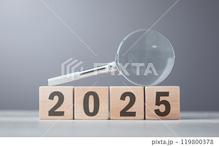 Wooden block displaying '2025' placed beside a magnifying glass on a table, set against a gray background. Symbolizes a closer look at future plans, analysis, and preparation for the year ahead 119800378