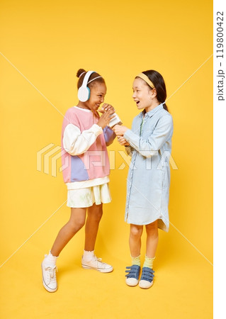 Vertical shot of smiling little Black girl in headphones singing in microphone with female friend wearing colorful casual clothes while having fun in yellow studio, copy space 119800422