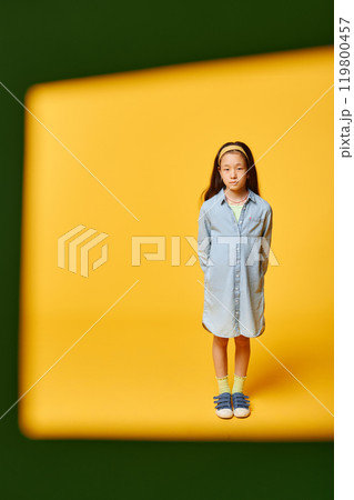 Vertical shot through green frame of cute preteen girl wearing denim shirt dress while standing against yellow background in studio, copy space 119800457