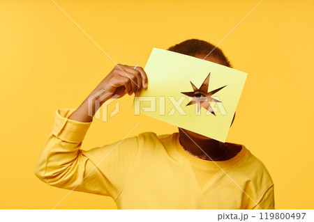 Little Black girl holding in hand paper sheet with cut out star while covering face looking through geometric shape at camera against yellow background in studio, copy space 119800497