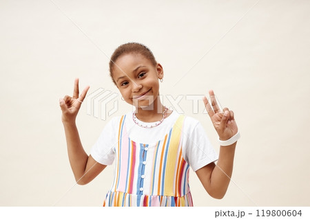 Medium portrait shot of cute young Black girl showing V sign with both hands wearing colorful dress and beaded choker smiling at camera while posing against neutral background in studio, copy space Medium portrait shot of cute young Black girl showing V sign with both hands wearing colorful dress and beaded choker smiling at camera while posing against neutral background in studio, copy space 119800604