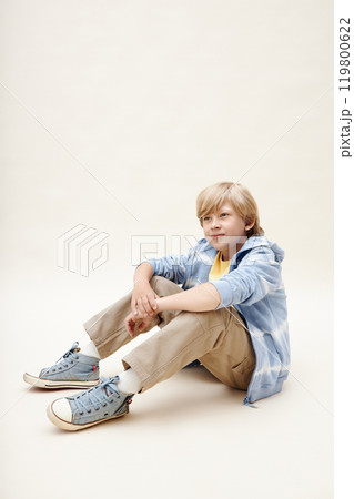 Vertical shot of preteen boy wearing blue sneakers and zip hoodie looking away while sitting on floor posing in studio against neutral background, copy space 119800622