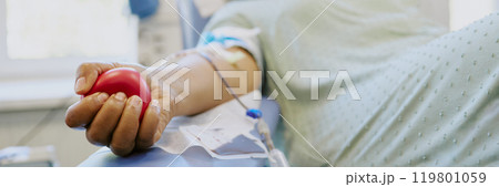 Close-up of a person donating blood at a medical clinic, holding a red squeeze ball. Intravenous equipment and hospital gown visible Close-up of a person donating blood at a medical clinic, holding a red squeeze ball. Intravenous equipment and hospital gown visible 119801059