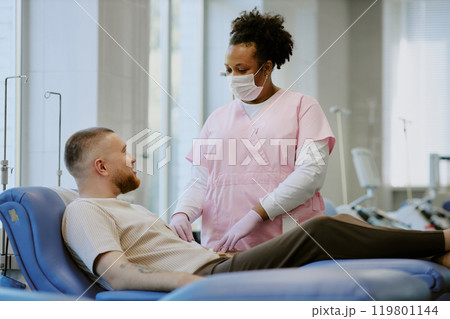 Medical professional in mask attending reclining patient in modern clinic setting with windows. Medical equipment visible in background creating professional healthcare environment 119801144