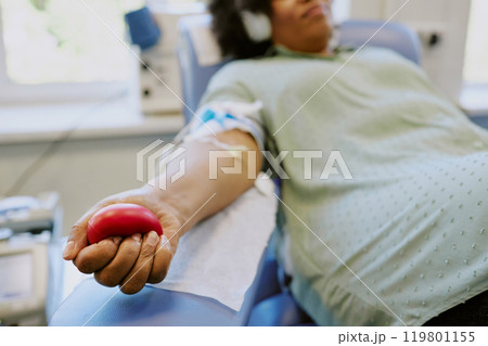 Close-up of person donating blood while squeezing a red ball. Patient in medical chair with intravenous line inserted, focusing on health and well-being Close-up of person donating blood while squeezing a red ball. Patient in medical chair with intravenous line inserted, focusing on health and well-being 119801155
