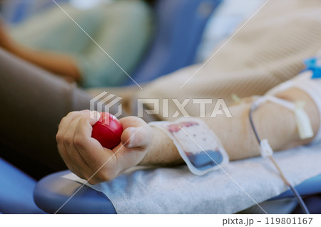 Arm holding red stress ball while donating blood in medical setting. Person is lying on hospital bed with IV line inserted into arm for procedure 119801167