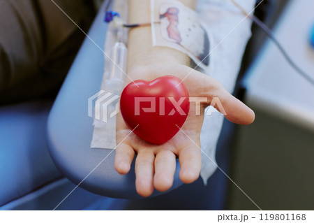 Close-up image showing person holding heart-shaped stress ball while donating blood. IV line visible on arm with background slightly blurred, emphasizing action 119801168