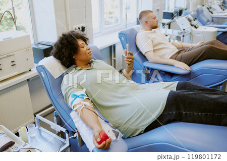 People reclined donating blood in medical facility, holding stress balls in arms Relaxing on chairs with medical equipment surrounding them, giving blood in safe and sterile environment People reclined donating blood in medical facility, holding stress balls in arms Relaxing on chairs with medical equipment surrounding them, giving blood in safe and sterile environment 119801172