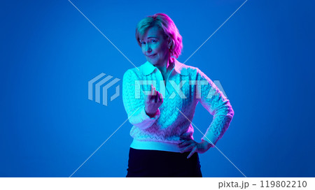 Elderly woman looking directly at camera, gesturing against blue studio background in neon light 119802210