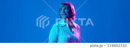 Portrait of beautiful elderly woman, smiling, looking upwards with dreamy face against blue studio background in neon light 119802332