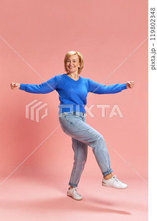 Full-length. Smiling, cheerful mature, elderly woman in jeans and sweater in motion, dancing against pink studio background 119802348