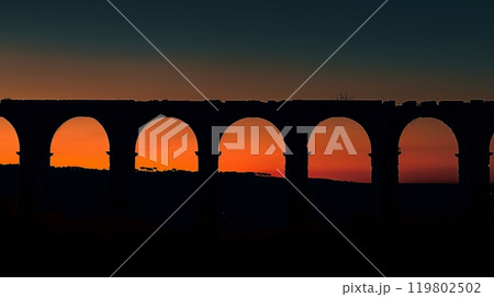 Ancient Roman aqueduct arches silhouetted against a glowing twilight sky, highlighted by the setting sun. 119802502