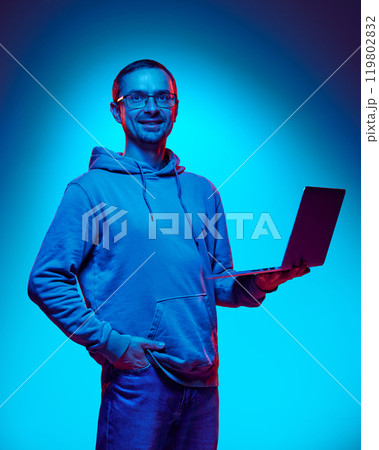 Young man, IT specialist looks confidently and smiling at camera holding laptop in neon light against gradient studio background. 119802832